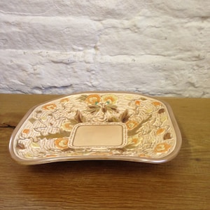 May include: A beige ceramic dish with a floral pattern in orange, yellow, and brown. The dish has a rectangular space in the centre and a curved edge. The dish is on a wooden surface.