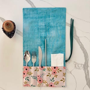 May include: A teal blue and floral print cutlery roll with a spoon, fork, knife, straw, and napkin. The roll is made of fabric and has a green tie closure. The floral print features pink flowers and green leaves.