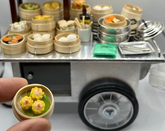 Può includere: Un carrello per il cibo in miniatura con numerosi piccoli cestini per la cottura a vapore in bambù pieni di vari ravioli. Un singolo cestino in bambù contenente tre ravioli con guarnizione gialla è tenuto in primo piano, separato dal carrello.