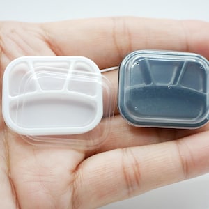 May include: Two small, divided food containers. One is white with a clear lid, the other is dark grey with a clear lid. Each container has three compartments. The containers are held in a person's hand.