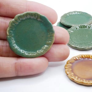 Puede incluir: Pequeños platos de cerámica en tonos verdes y marrones. Los platos tienen un diseño de borde festoneado. Un plato se sostiene en una mano, con varios otros visibles en el fondo. Estos pequeños platos son probablemente para casas de muñecas o exhibiciones en miniatura.
