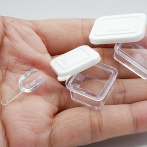 Könnte beinhalten: Eine Sammlung kleiner, quadratischer Behälter aus transparentem Kunststoff mit weißen Deckeln und einem kleinen Löffel. Die Behälter sind für die Aufnahme kleiner Gegenstände konzipiert. Die Gegenstände werden in einer Hand gehalten.