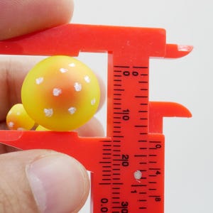 May include: A close-up of a yellow and orange mushroom figurine with white spots, being measured by a red caliper. The caliper is showing the mushroom's size. The background is plain white.