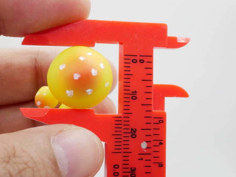 May include: A close-up of a yellow and orange mushroom figurine with white spots, being measured by a red caliper. The caliper is showing the mushroom's size. The background is plain white.