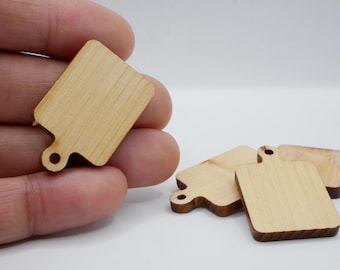 May include: A collection of small, wooden cutting boards. One is held in a hand, while several others are arranged on a white surface. The boards are rectangular with a handle and a hole for hanging. The wood grain is visible.