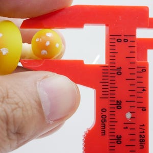 May include: Close-up of two miniature mushroom figurines, one larger than the other, held by a red caliper. The mushrooms are yellow with orange shading and white spots. The caliper is marked with measurements in inches.