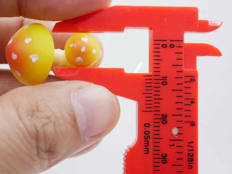 May include: Close-up of two miniature mushroom figurines, one larger than the other, held by a red caliper. The mushrooms are yellow with orange shading and white spots. The caliper is marked with measurements in inches.
