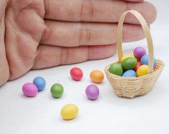 Può includere: Un piccolo cestino di vimini pieno di uova di Pasqua colorate. Il cestino ha un manico di legno e si trova su una superficie bianca. Ci sono anche diverse uova colorate sparse intorno al cestino.