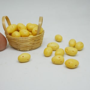 Puede incluir: Una pequeña cesta de mimbre llena de patatas amarillas. Varias patatas adicionales están esparcidas alrededor de la cesta sobre una superficie blanca.
