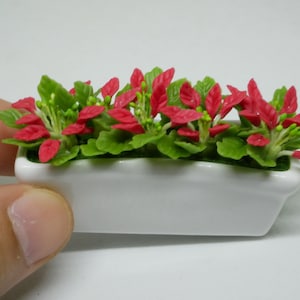 以下が含まれることがあります： 赤と緑の花が咲いた、ミニチュアの白いセラミック製のプランター。プランターは長方形で、わずかに湾曲した上部があります。