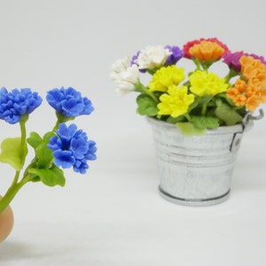 May include: A small bouquet of blue flowers in a hand, with a larger bouquet of multi-colored flowers in a metal bucket in the background.