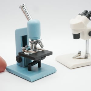 Può includere: Due microscopi giocattolo, uno blu e uno bianco, su una superficie bianca. Il microscopio blu ha un braccio argentato e un tavolino nero. Il microscopio bianco ha un braccio argentato e una base nera.