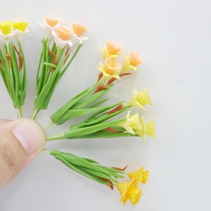 May include: A bouquet of yellow and orange daffodils with green stems. The flowers are made of polymer clay and are arranged in a fan shape.
