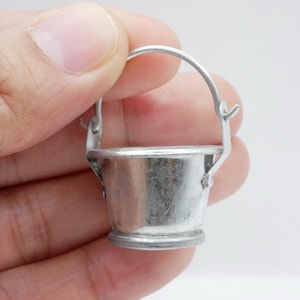 May include: A miniature silver metal bucket with a curved handle. The bucket has a rounded base and a simple design. The handle is attached with small hinges. The bucket is held in a hand. The bucket is likely a decorative item.