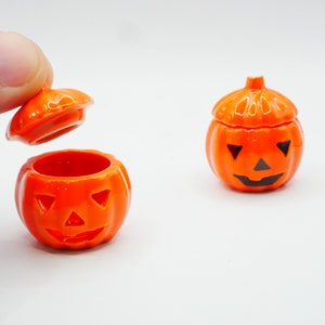 May include: Two orange ceramic pumpkin-shaped containers with black jack-o'-lantern faces. One container is open, revealing the inside.