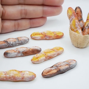 May include: A collection of miniature, hand-crafted bread loaves. The loaves are in a small wicker basket and on a white surface. Some loaves are brown and some are yellow.