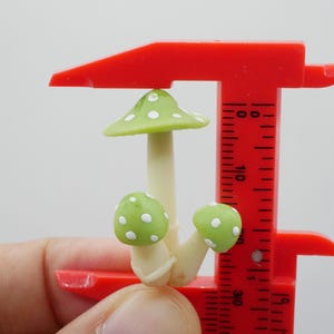 May include: A miniature mushroom sculpture with a light green cap and white polka dots, held by a red caliper. The mushroom has a cream-colored stem and two smaller green mushrooms with white dots. The caliper is marked with measurements.