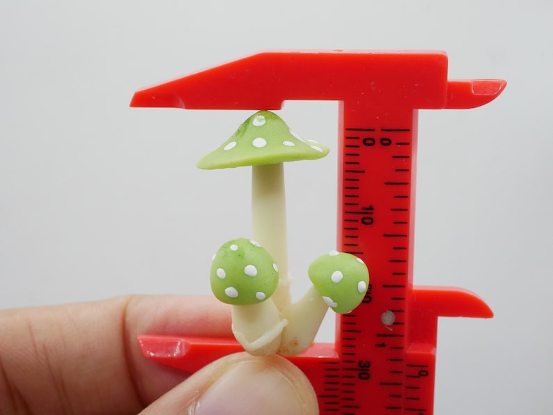 May include: A miniature mushroom sculpture with a light green cap and white polka dots, held by a red caliper. The mushroom has a cream-colored stem and two smaller green mushrooms with white dots. The caliper is marked with measurements.
