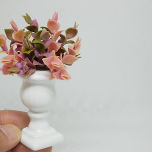 May include: A miniature white ceramic planter with a square base and a round body, holding a bouquet of pink, green, and purple artificial flowers.