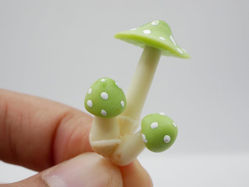 May include: A miniature mushroom sculpture with a light green cap and white polka dots. The stem is off-white. The sculpture is a whimsical decorative item.