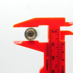 Pu&ograve; includere: Un calibro a corsoio rosso in plastica misura un piccolo oggetto rotondo di colore dorato. Il calibro ha un righello nero con segni in pollici e frazioni di pollice.