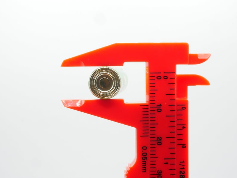 Pu&ograve; includere: Un calibro a corsoio rosso in plastica misura un piccolo oggetto rotondo di colore dorato. Il calibro ha un righello nero con segni in pollici e frazioni di pollice.
