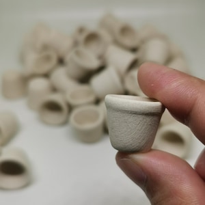 May include: A close-up of a small, white ceramic pot. The pot is being held in a hand, and there are many other similar pots in the background.
