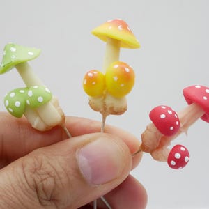 May include: Three miniature mushroom figurines with colorful caps and white polka dots. The mushrooms have yellow, red, and green caps. The stems are white and beige. The figurines are held in a hand against a white background.