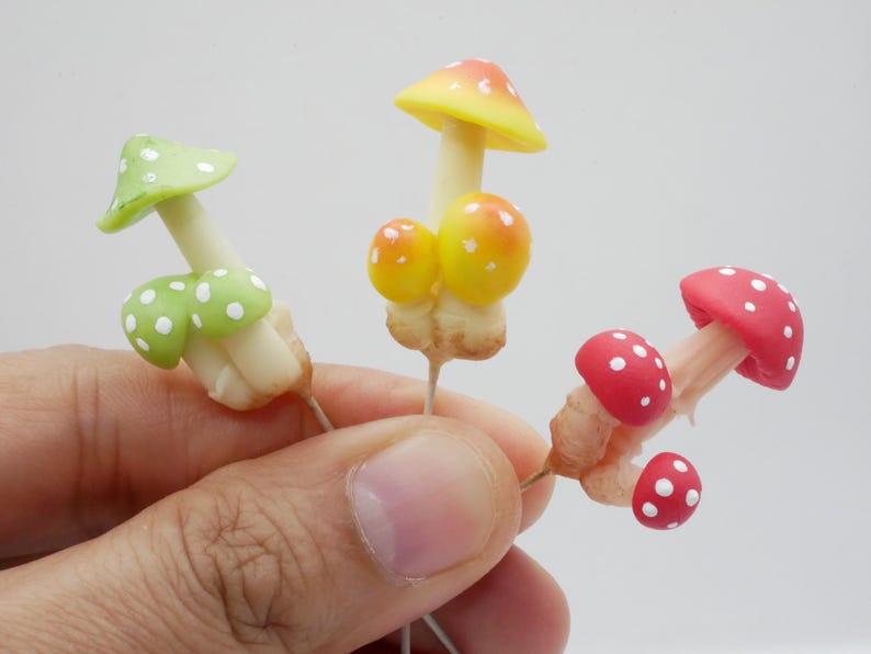 May include: Three miniature mushroom figurines with colorful caps and white polka dots. The mushrooms have yellow, red, and green caps. The stems are white and beige. The figurines are held in a hand against a white background.