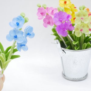 May include: A miniature arrangement of colorful orchid flowers. A single stem of light blue orchids with green leaves is held in a hand. A small silver bucket holds a variety of orchids in pink, purple, yellow, and green.