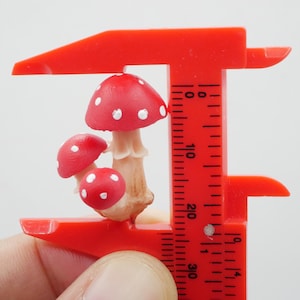 May include: A miniature mushroom figurine with red caps and white spots is held by a red caliper. The mushrooms have beige stems. The caliper is marked with measurements in inches.