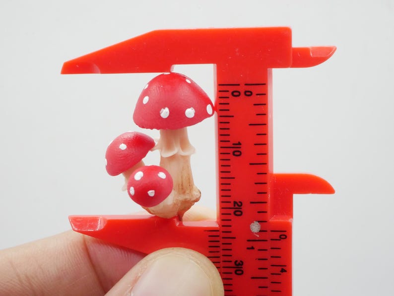 May include: A miniature mushroom figurine with red caps and white spots is held by a red caliper. The mushrooms have beige stems. The caliper is marked with measurements in inches.
