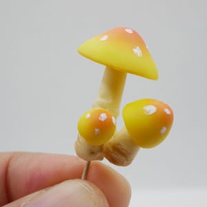 May include: A cluster of three miniature mushrooms. The largest mushroom has a yellow cap with orange tips and white spots. The smaller mushrooms have yellow caps with orange spots. The stems are beige.