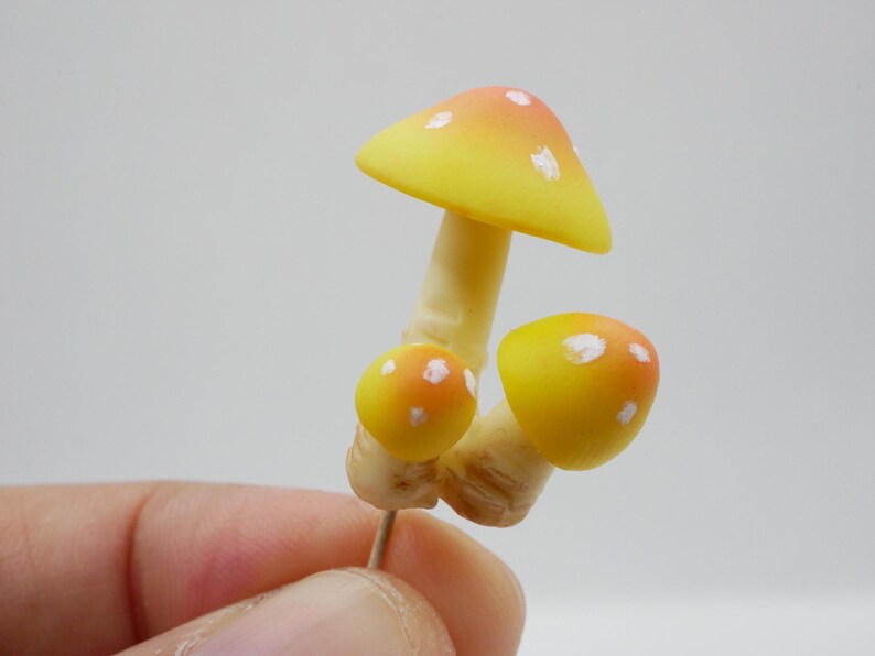 May include: A cluster of three miniature mushrooms. The largest mushroom has a yellow cap with orange tips and white spots. The smaller mushrooms have yellow caps with orange spots. The stems are beige.