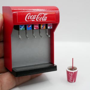 Peut inclure: Miniature fontaine à soda Coca-Cola rouge avec le logo Coca-Cola. La fontaine propose plusieurs options de soda et un petit gobelet de soda avec une paille. La fontaine est rouge et argentée, et le gobelet est rouge et blanc.