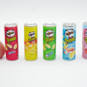 Könnte beinhalten: Sechs Mini-Dosen Pringles Kartoffelchips in verschiedenen Geschmacksrichtungen: Original, Sauerrahm & Zwiebel, Cheddar & Sauerrahm, Spicy Chicken und Salz & Essig. Die Dosen sind in einer Reihe auf einer weißen Oberfläche angeordnet.