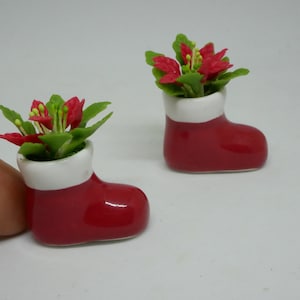 May include: Two miniature red ceramic boots with white trim, each holding a small potted poinsettia plant with red flowers and green leaves.