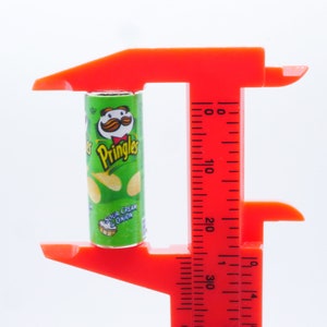 Pu&ograve; includere: Uno strumento di misurazione in plastica rossa con un contenitore di patatine Pringles verde e giallo all'interno. La lattina &egrave; etichettata "Pringles Sour Cream & Onion".