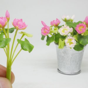 以下が含まれることがあります： ピンクと白の人工蓮の花の2つのアレンジ。1つのアレンジは小さな金属製のバケツに入っていて、もう1つは葉のついた単一の茎です。