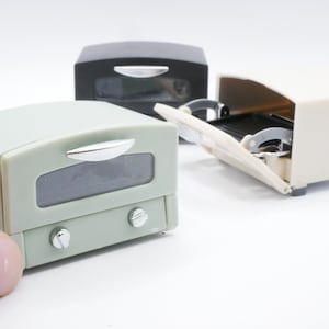 以下が含まれることがあります： 緑、白、黒の3色のミニチュアトースターオーブン。緑のトースターオーブンは手前にあり、クロム製のハンドルが付いている。白いトースターオーブンは開いており、内部が見える。黒いトースターオーブンは閉じている。