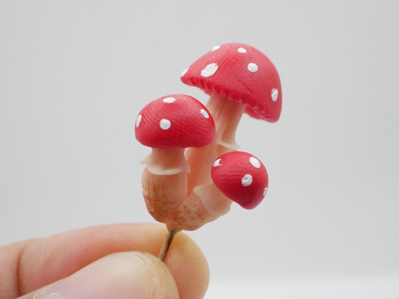 May include: A cluster of three miniature mushrooms with bright red caps and white spots. The stems are a pale beige color. The mushrooms are held by a hand against a plain white background. The overall theme is nature and fantasy.