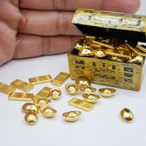 Puede incluir: Un pequeño cofre del tesoro pintado de oro, abierto para revelar un montón de lingotes y monedas de oro. El cofre tiene detalles en negro, y las piezas de oro están esparcidas a su alrededor. La imagen sugiere temas de riqueza y fantasía, con un enfoque en miniaturas coleccionables.