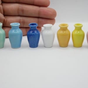 May include: Seven miniature ceramic vases in pastel shades of mint green, light blue, dark blue, white, yellow, orange, and pink. The vases are displayed on a white surface.