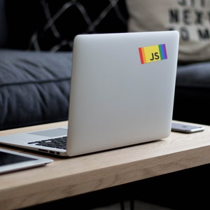 K&ouml;nnte beinhalten: Ein silberner Laptop mit einem Regenbogen- und gelben Aufkleber mit der Aufschrift "JS" auf der R&uuml;ckseite. Der Laptop steht auf einem Holztisch mit einem Tablet und einem Telefon.