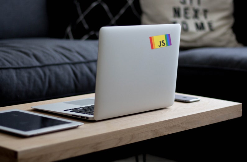 K&ouml;nnte beinhalten: Ein silberner Laptop mit einem Regenbogen- und gelben Aufkleber mit der Aufschrift "JS" auf der R&uuml;ckseite. Der Laptop steht auf einem Holztisch mit einem Tablet und einem Telefon.