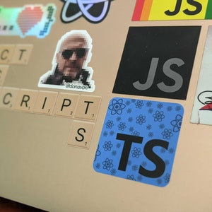 May include: A laptop covered in stickers, including a pixelated image of a person, the text "JS", and a blue sticker with "TS". Scrabble tiles spell out "REACT" and "JAVASCRIPT".