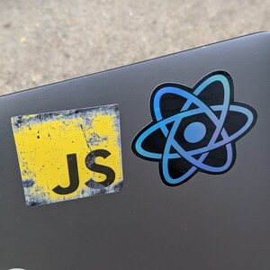 May include: A grey laptop with two stickers on it. One sticker is yellow with the text "Js" on it. The other sticker is black with a blue and purple atom symbol.