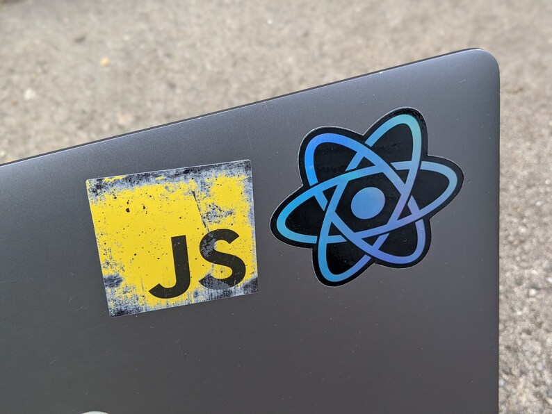 May include: A grey laptop with two stickers on it. One sticker is yellow with the text "Js" on it. The other sticker is black with a blue and purple atom symbol.