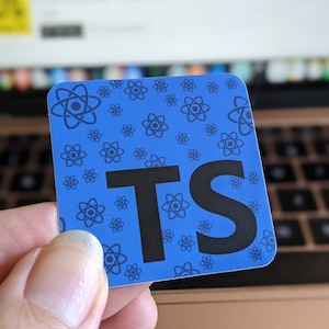 May include: A blue square sticker with the letters "TS" in black. The sticker has a pattern of small atom symbols in a darker shade of blue. The sticker is held in front of a laptop keyboard.
