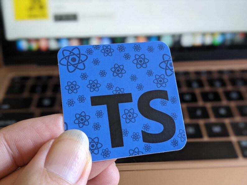 May include: A blue square sticker with the letters "TS" in black. The sticker has a pattern of small atom symbols in a darker shade of blue. The sticker is held in front of a laptop keyboard.